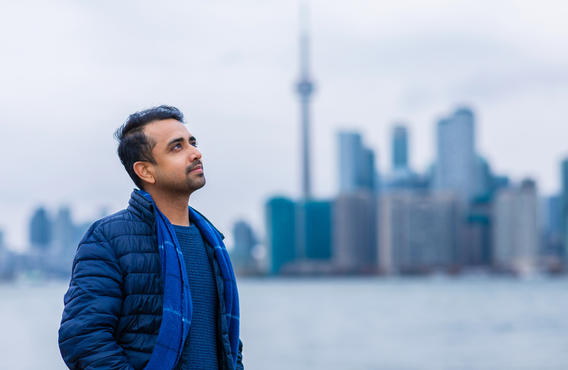 Un homme debout près d’un port de Toronto, regardant au loin, les mains dans les poches. Derrière lui se trouve le paysage du centre-ville de Toronto avec la tour CN visible.