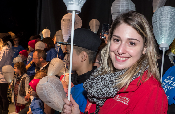 At a Light The Night event, A woman with dirty blonde hair is smiling while holding up a white lantern. A crowd of people are around her and there's a person on a microphone not far from her on the left side of the image.